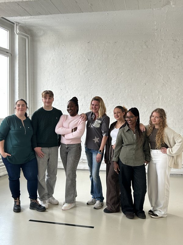 7 Menschen stehen vor einer weißen Wand und schauen in die Kamera