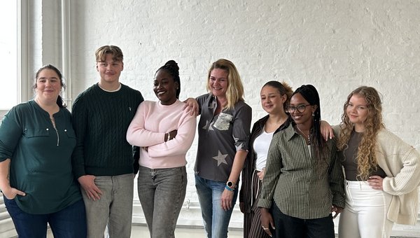 7 Menschen stehen vor einer weißen Wand und schauen in die Kamera