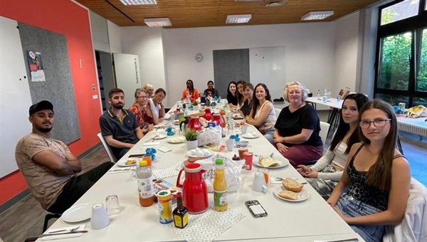 Personen sitzen an einem langen Tisch in einem Raum. Auf dem Tisch stehen Getränke wie Saft und Kaffee sowie Lebensmittel wie Brötchen und Marmelade. Der Raum hat weiße Wände, eine Holzdecke mit eingebauten Leuchten und große Fenster auf der rechten Seite. Die linke Wand ist rot. An der linken Wand befindet sich ein Whiteboard.